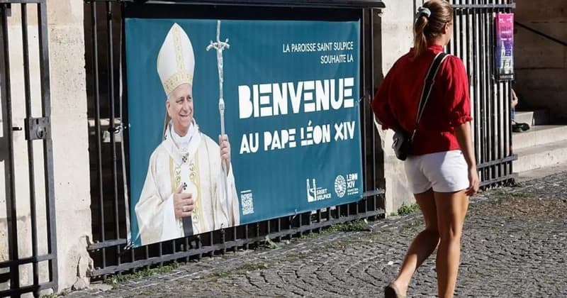 A promotional banner at the Saint-Sulpice parish in Paris welcomes 'Pope Leo XIV', featuring a portrait of a man in papal attire holding a ferula. A woman wearing a red shirt and white shorts is captured mid-stride as she walks past the black iron gate where the banner is displayed on a cobblestone street.