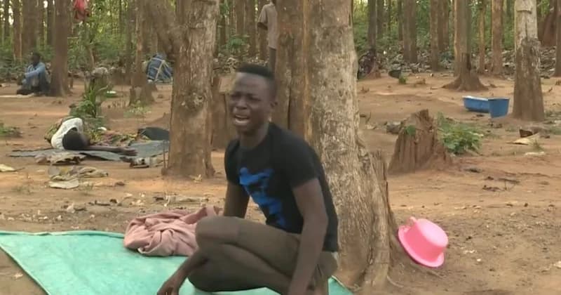 A young man with an emotional expression sits on a green mat in a wooded outdoor area, likely a prayer camp. In the background, other individuals are seen resting on the ground among trees and various personal items like plastic basins and cloths.