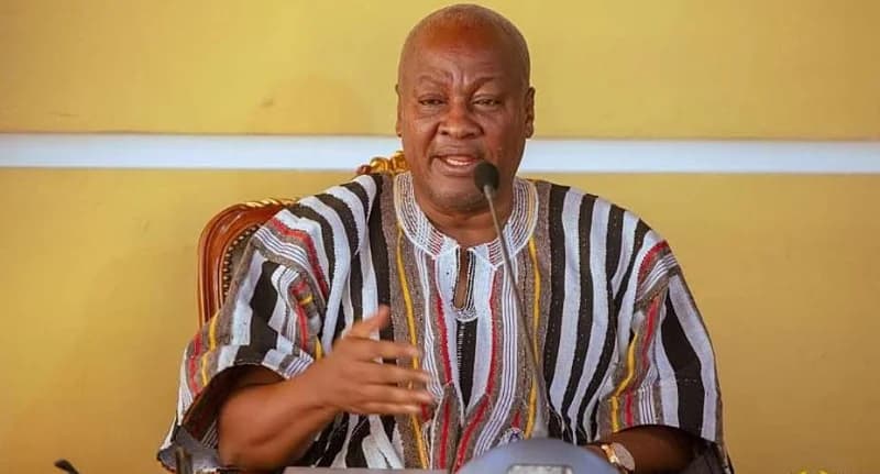 President John Dramani Mahama is captured speaking at an event, gesturing with his hand while seated in an ornate chair. He is wearing a traditional Ghanaian smock (batakari) with black, white, and red stripes, positioned behind a microphone against a solid yellow background.
