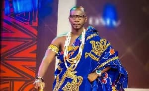 Ghanaian musician Okyeame Kwame is pictured in a studio setting wearing regal blue and gold traditional cloth. He is adorned with several layers of gold bead necklaces and armbands, holding a traditional staff while looking confidently at the camera.