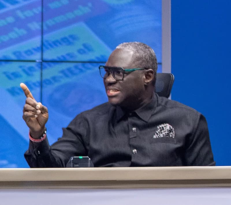 Palgrave Boakye Danquah, a government spokesperson, is captured in a studio setting, gesturing with his finger while speaking emphatically. He is wearing a black collared shirt, glasses, and a patterned pocket square against a blue background with a blurred news screen.