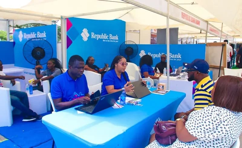 Staff members of Republic Bank Ghana engage with customers at a branded exhibition booth during the JoyNews Habitat Fair. The image captures several bank representatives using laptops to assist visitors under a white outdoor tent setup with prominent blue branding.