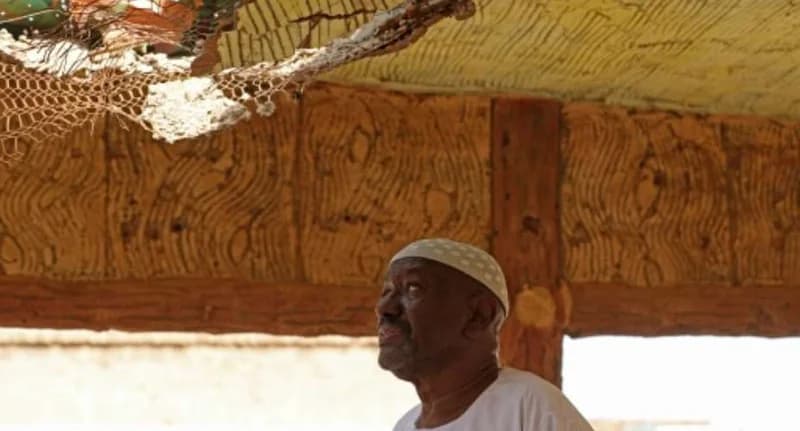 An elderly man wearing a white skullcap looks up at the extensive damage to the ceiling of a building. The structure shows a large hole with exposed wire mesh and crumbling plaster, illustrating the wreckage faced by those returning to Sudan's capital.