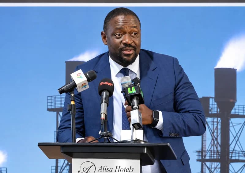 A man in a navy blue suit speaks at a podium during a presentation at Alisa Hotels. Behind him, a screen displays an image of industrial gas flaring towers, likely illustrating the discussion on methane emissions and energy infrastructure in Ghana.