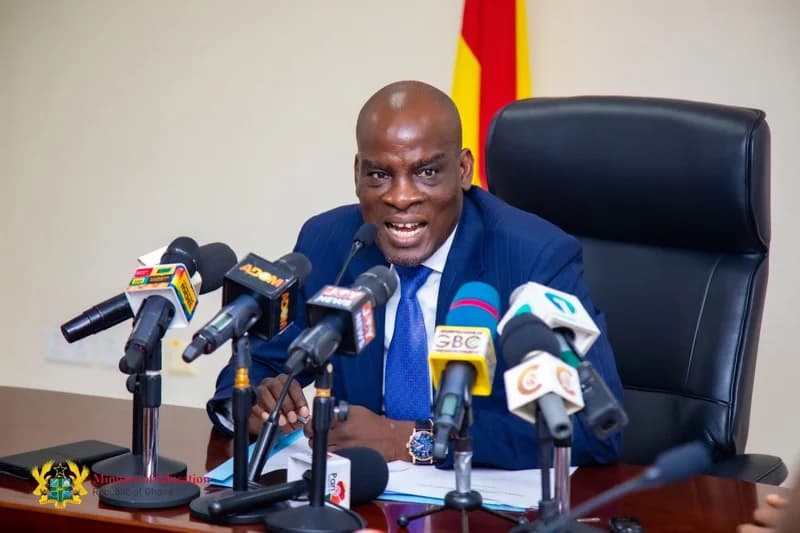 A man in a blue pinstripe suit speaks passionately during a press conference surrounded by numerous microphones from various Ghanaian media outlets. The scene includes a glimpse of the Ghana national flag in the background and a watermark for the Ministry of Education, Republic of Ghana.