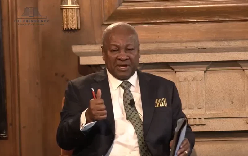 President John Dramani Mahama is pictured speaking during a formal event, gesturing with his hand while holding a pen. He is dressed in a professional dark suit with a patterned green tie, positioned against a background of ornate wood paneling that includes a watermark of the Republic of Ghana's presidency.