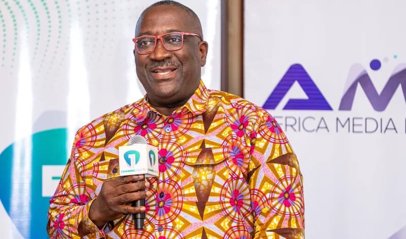 Samuel Attah-Mensah, a prominent Ghanaian media executive, is pictured speaking into a Channel One TV microphone. He is wearing a vibrant, patterned African print shirt and red-framed glasses while standing against a backdrop featuring media branding.