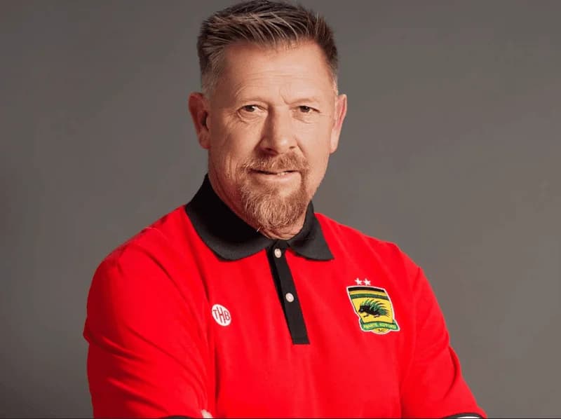 A professional portrait of Eric Tinkler, the former coach of Asante Kotoko, wearing the club's official red polo shirt with a black collar. He is featured against a solid grey background, looking directly at the camera with a slight smile.