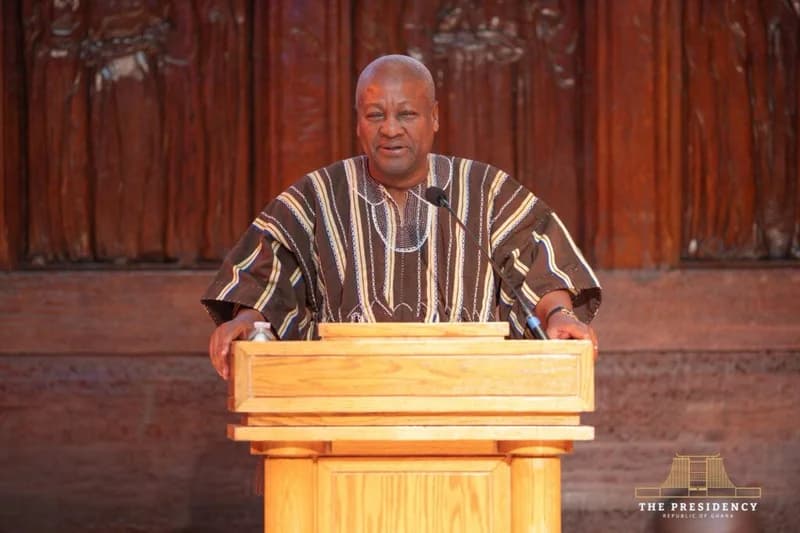 President John Dramani Mahama is captured speaking from a wooden lectern during an official address. He is dressed in a traditional striped Ghanaian smock against a background of intricate wood carvings, featuring the official logo of the Presidency of the Republic of Ghana.