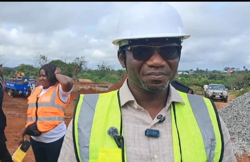 A man, likely the Bono East Regional Minister, wearing a white hard hat and a lime-green high-visibility vest, stands at a construction site. In the background, a woman in an orange safety vest and construction vehicles are visible on the red earth terrain of the St. Dominic College project site in Tuobodom.