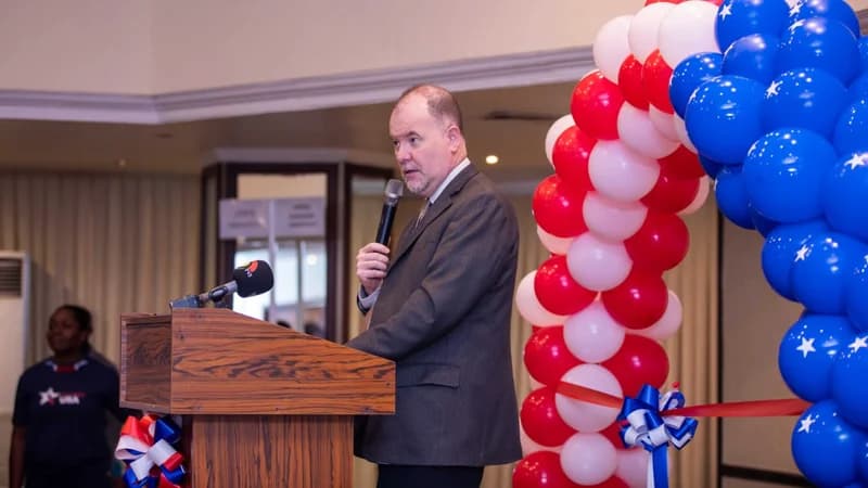 A male official in a grey suit delivers a speech at a wooden lectern during a US-themed event. Beside him is a decorative arch of red, white, and blue balloons, and a person in a 'USA' branded shirt is visible in the background. The setting suggests an official ceremony or announcement by the US Embassy in Ghana.