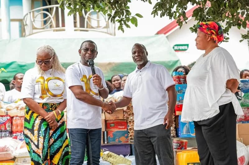 Representatives from RCI Immanuel Temple are seen shaking hands while presenting a large donation of food and household supplies to the Motherly Love Orphanage. The donors are wearing white t-shirts commemorating their 10th anniversary, surrounded by stacks of boxed goods, sacks of grain, and bottled beverages.