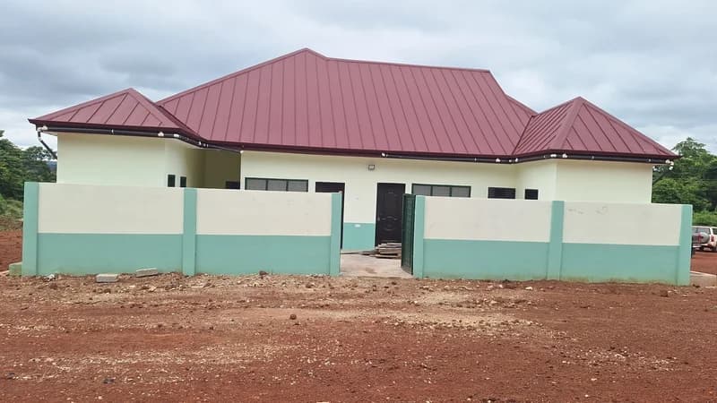 A newly constructed health facility building, identified as a CHPS Compound in Kokoago, Bono East. The structure features a prominent red metal roof and a light green and white perimeter wall, situated on reddish earth under a cloudy sky.