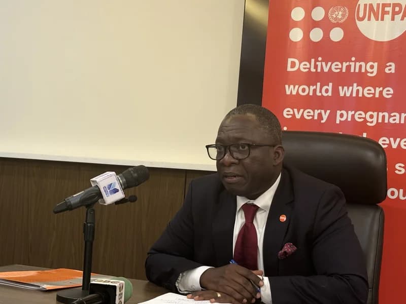 A man in a dark suit and red tie sits at a desk during a press conference or official meeting in Ghana. Behind him is a prominent red UNFPA banner with text about maternal health, while several microphones from various news outlets are positioned in front of him.