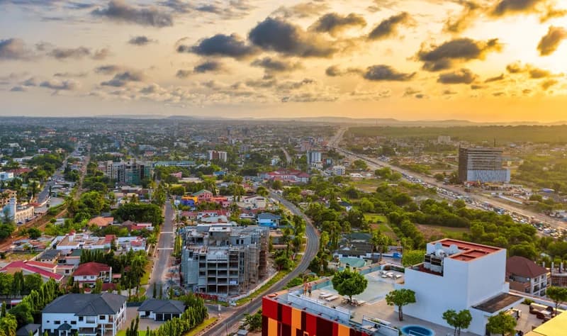 An expansive aerial view of an urban landscape in Ghana, likely Accra, during sunset or sunrise. The image features a mix of residential housing, modern multi-story developments under construction, and a busy highway stretching into the distance under a warm, cloud-filled sky.