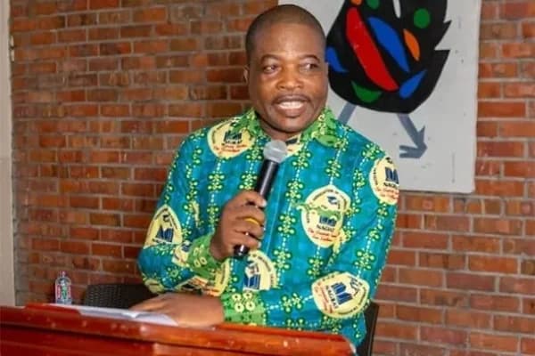 Angel Carbonu, President of the National Association of Graduate Teachers (NAGRAT), is seen speaking into a microphone at a podium. He is wearing a distinctive green and yellow shirt featuring the NAGRAT logo, set against a background of a red brick wall and colorful artwork.