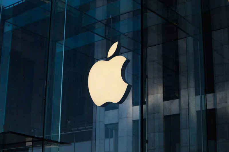 A close-up photograph of a glowing white Apple logo displayed behind a modern glass facade of a building. The surrounding architecture features dark blue-tinted glass and stone pillars, creating a sleek, corporate aesthetic.