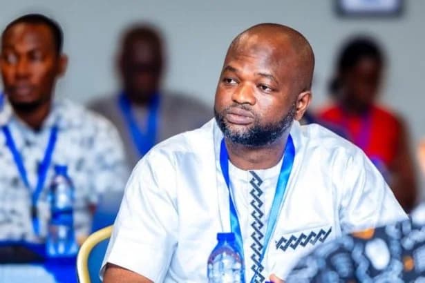 A photograph of Kwasi Kwarteng, the Public Relations Officer for the Ministry of Education in Ghana, at an official event. He is wearing a white traditional-style shirt with blue accents and a blue lanyard, while other attendees are seen blurred in the background.