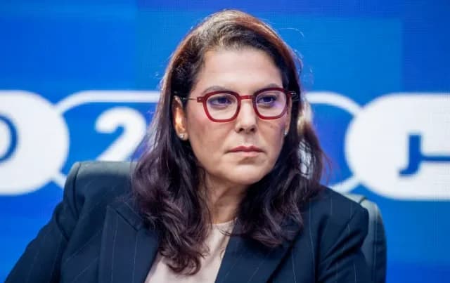 A medium shot of Fatima Zahra El Mansouri, who has been appointed as Morocco's first female Prime Minister. She is pictured wearing distinctive red-rimmed glasses and a dark pinstripe blazer against a blue backdrop with blurred text.