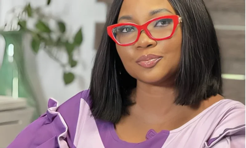 A close-up portrait of actress Jumoke Odetola, who is seen wearing bright red-framed glasses and a purple patterned outfit. She has a subtle smile and is looking directly at the camera in an indoor setting with a soft-focus background.