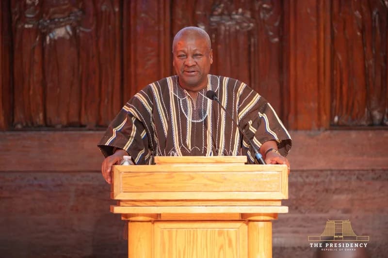 President John Dramani Mahama is seen delivering a speech from a wooden podium. He is dressed in a traditional brown and white striped northern smock, known as a fugu, against a background of detailed wood carvings.