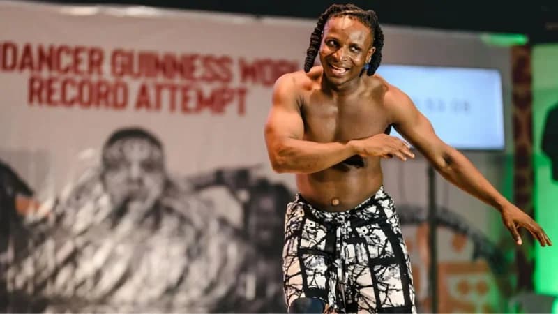 A shirtless man with dreadlocks, identified as Ben Dancer, is captured mid-dance during his Guinness World Record attempt. In the background, a large banner displays text related to the record attempt, and a digital timer is visible on a monitor.