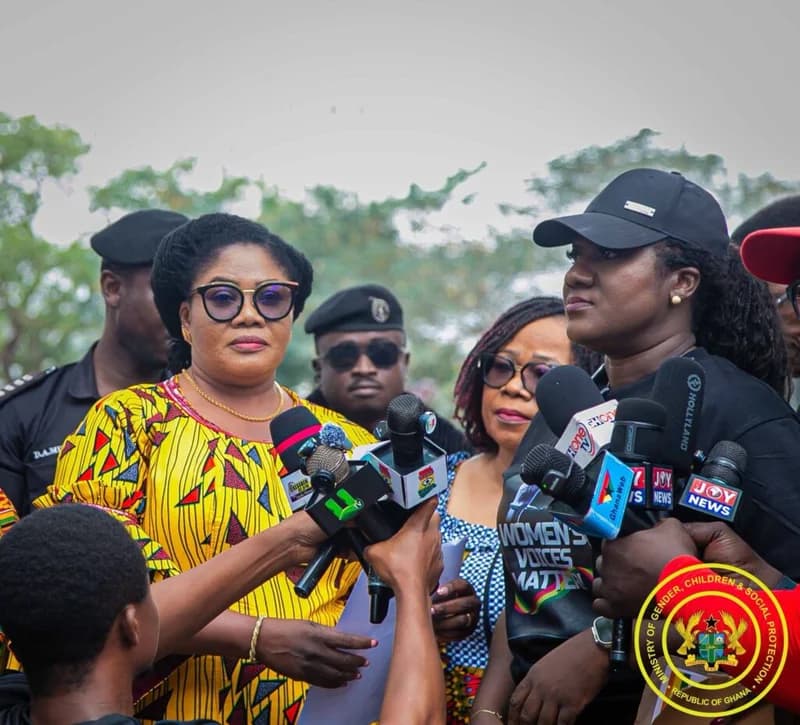 A woman in a vibrant yellow and black patterned dress, likely a Ghanaian government official, addresses the media while surrounded by numerous microphones. She is accompanied by a woman in a black shirt that reads 'WOMEN'S VOICES MATTER', with security personnel and journalists visible in the background.