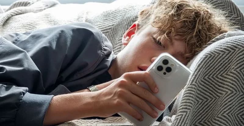 A young teenager with curly hair is lying on a bed while focused on a white smartphone. The image captures a common scene of youth engagement with digital technology in a personal space.