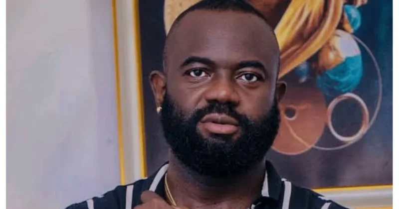 A close-up portrait of late Nollywood actor CJay Nwoks, featuring his characteristic full black beard and short hair. He is wearing a black and white striped shirt and stands in front of a colorful framed painting.