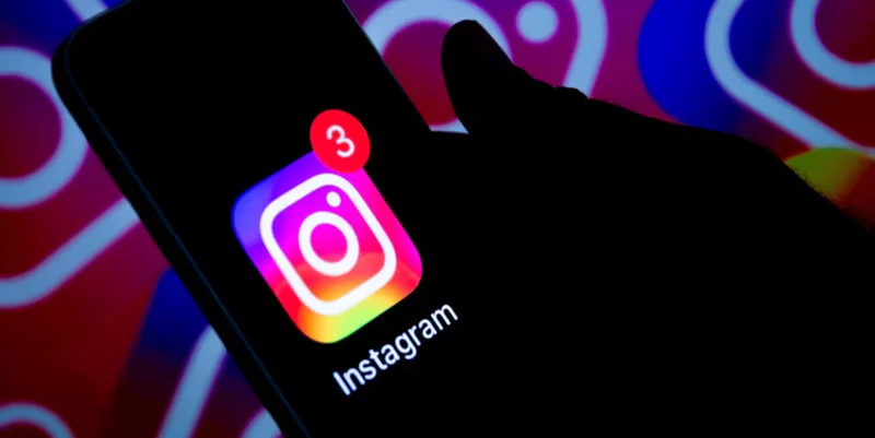 A close-up photograph of a hand holding a smartphone in low light, displaying the Instagram app icon with a notification badge showing the number three. The background is illuminated with a stylized, colorful projection of the Instagram logo's camera outline and gradient.
