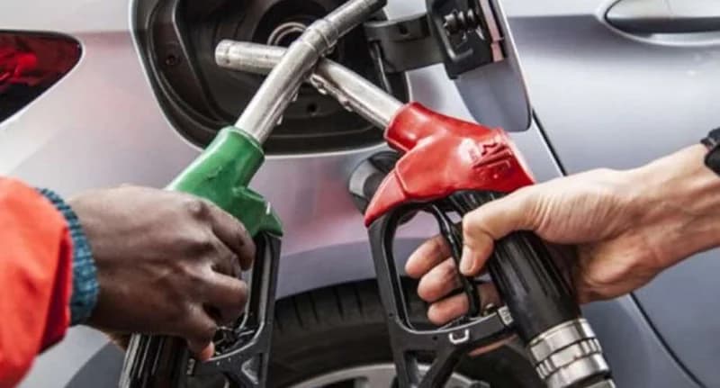 A close-up photograph showing two fuel pump nozzles, one red and one green, being held by two different individuals near the open fuel tank of a silver vehicle. The image captures the moment of refueling at a service station, emphasizing the energy sector.