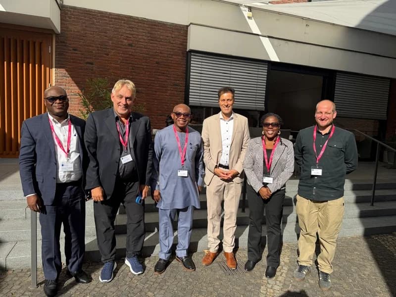 A group of six academics and officials from KNUST and the University of Bonn posing for a group photograph outdoors. Most of the individuals are wearing identification lanyards, and they are standing in front of a modern building with brick and glass architectural elements. This meeting highlights the ongoing collaboration between the two institutions to launch a joint PhD programme.