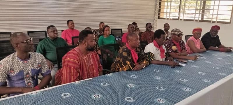 A group of individuals, likely members of the Technical University Teachers' Association of Ghana (TUTAG), are seated at a long table during a meeting or press conference. Several people are wearing red scarves or clothing, which is a traditional sign of industrial action or protest in Ghana. The meeting takes place in a room with white paneled walls and a patterned blue tablecloth.
