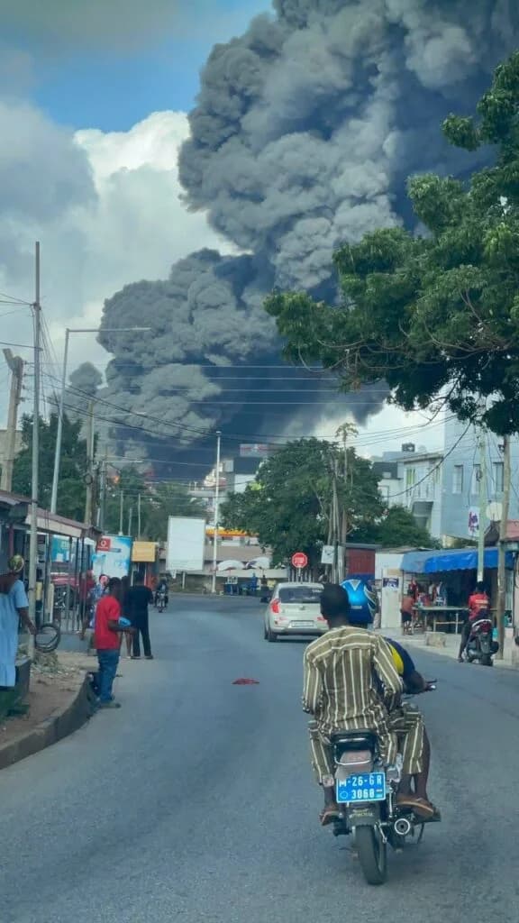 A massive plume of thick, dark grey smoke billows into the sky over an urban street in Accra. Onlookers and commuters on motorcycles have stopped to witness the large-scale fire occurring in the distance, likely at the Duraplast factory in the North Industrial Area.