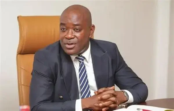 Professor Ahmed Jinapor Abdulai, the Director-General of the Ghana Tertiary Education Commission (GTEC), is pictured seated at a desk in a professional setting. He is dressed in a dark navy suit with a blue and white striped tie, with his hands clasped as if engaged in a discussion or presentation.