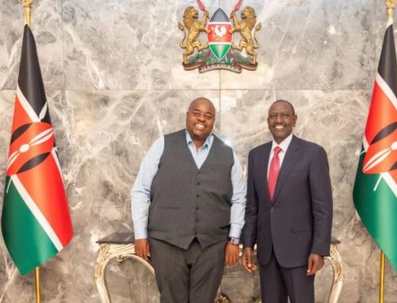 Zimbabwean businessman Wicknell Chivayo (left) and Kenyan President William Ruto (right) are pictured smiling together in an official setting. They are positioned in front of a marble wall featuring the Kenyan Coat of Arms and flanked by two Kenyan national flags.