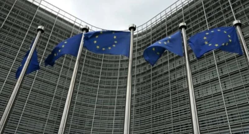 Several European Union flags flutter on tall silver flagpoles in front of the curved glass facade of the Berlaymont building in Brussels. The image represents the institutional heart of the EU during a period of high-level policy discussions.