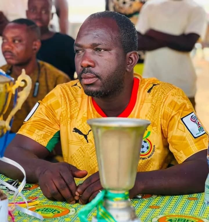 A man wearing a yellow Ghana national team football jersey is seated behind a table during a sports-related event. A silver trophy is placed prominently in the foreground, while other people and another gold-colored trophy are visible in the background.
