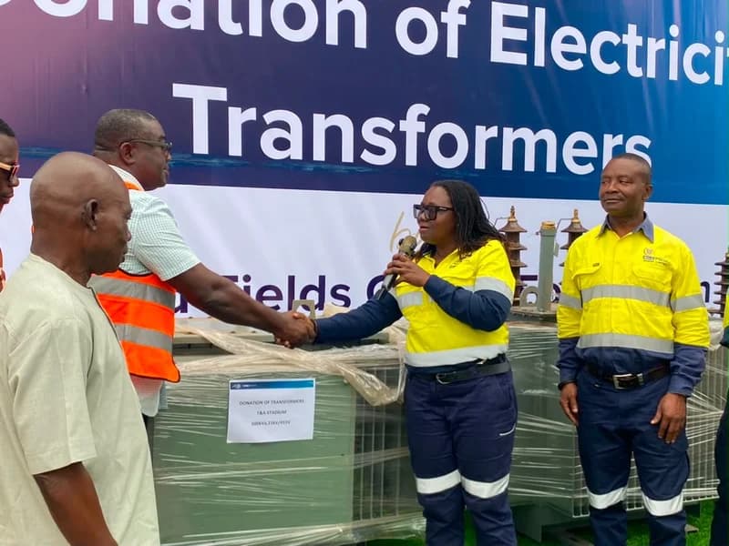 A representative from Gold Fields Ghana Limited shakes hands with an official during a ceremony for the donation of electricity transformers. The event features a large transformer wrapped in plastic in the foreground and a backdrop banner highlighting the donation for the T&A Stadium.