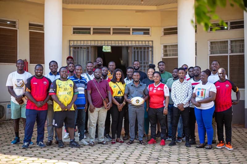 A large group of approximately 28 people, likely teachers and sports officials, pose for a photograph in a paved courtyard in front of a building with white pillars. A man in the center holds a white rugby ball, representing the Ashanti Rugby League during their review and planning session. The group is dressed in various casual and athletic outfits, including some wearing official team jerseys.