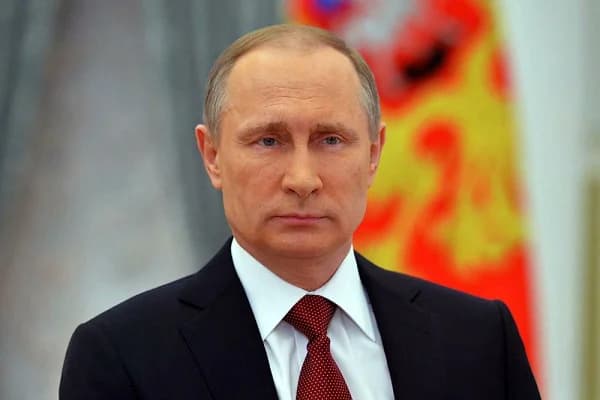 A close-up portrait of Russian President Vladimir Putin looking directly forward with a stern expression. He is wearing a formal dark suit, white shirt, and a patterned burgundy tie against a blurred background featuring the colors of the Russian state flag.