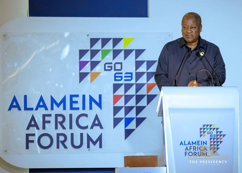 President John Dramani Mahama is pictured delivering a speech at a podium during the Alamein Africa Forum. He is dressed in a black shirt, standing before a large backdrop that displays the event's name and a geometric representation of the African continent.