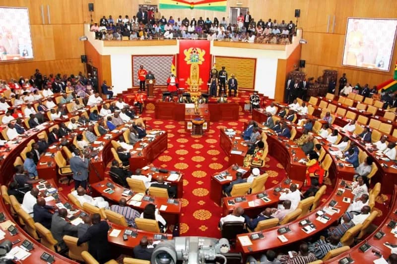 A wide-angle interior view of the Parliament of Ghana during a sitting. Members of Parliament are seated in a semi-circular arrangement facing the Speaker's dais, which features the national coat of arms against a red backdrop. A crowded public gallery is visible above the chamber floor.