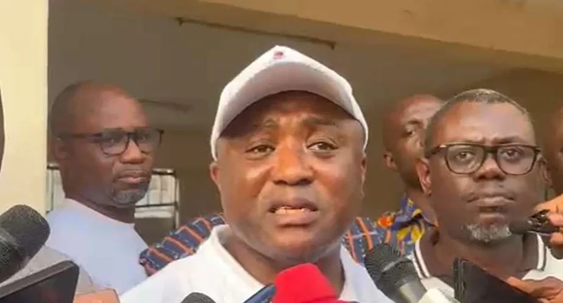 A man wearing a white cap and polo shirt addresses the media while surrounded by microphones and other officials. He appears to be speaking at a press briefing following negotiations between teacher unions and the government.