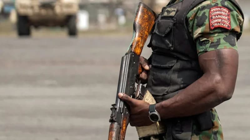 A member of a specialized military unit stands holding an assault rifle, wearing a camouflage uniform and a black tactical vest. A prominent red patch on the arm identifies the soldier as part of a Special Force Commando Ranger unit, likely in Nigeria.