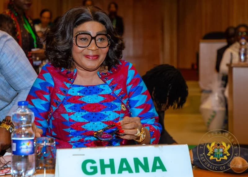 Former First Lady of Ghana, Lordina Mahama, is captured at a formal event seated behind a placard that reads 'GHANA'. She is wearing a vibrant blue and red patterned dress, large-framed glasses, and is holding a second pair of spectacles while looking toward the side.