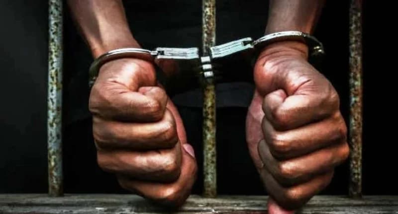 A conceptual photograph showing a person's hands in metal handcuffs held up against rusty iron bars. The image symbolizes arrest, imprisonment, or legal detention, fitting the context of a criminal investigation.