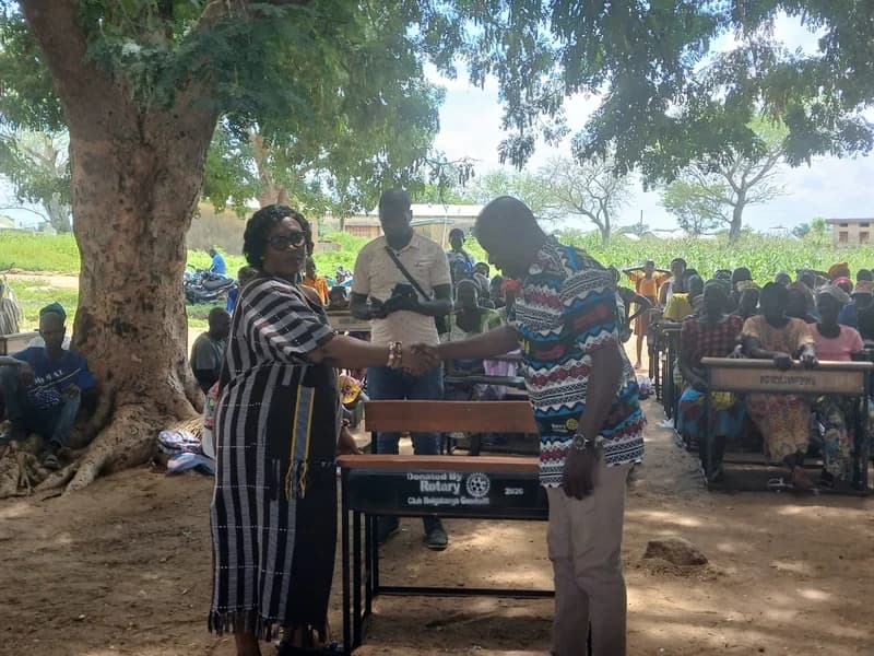 A woman and man shake hands over a newly donated dual school desk during an outdoor ceremony under a large tree in a rural setting. The desk features a label indicating it was donated by the Rotary Club of Bolgatanga Goodwill, with a large crowd of community members seated at similar desks in the background.