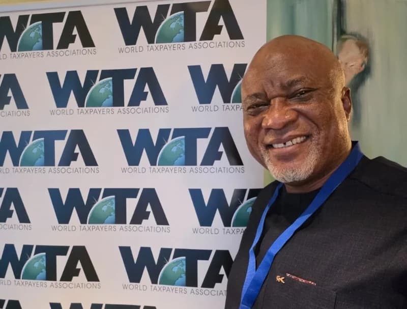 Ghanaian politician Hopeson Adorye is seen smiling in front of a backdrop for the World Taxpayers Associations (WTA). He is wearing a black traditional-style shirt with a blue lanyard around his neck, indicating his participation in an official event.