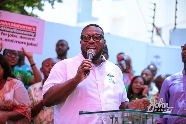 George Opare Addo, the National Youth Organizer of the National Democratic Congress (NDC), is captured speaking at a podium during a political event. He is holding a microphone and addressing a crowd, with supporters and campaign placards visible in the background.